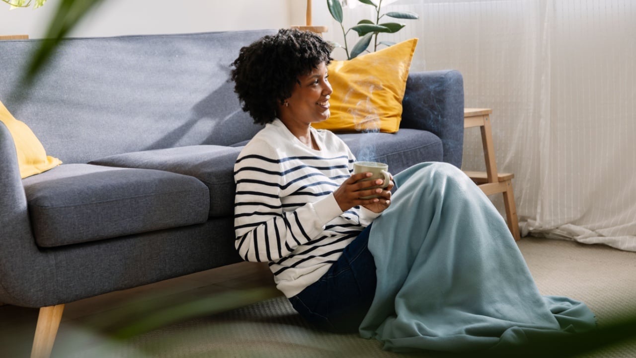 Young Latin American black woman relaxing on sofa drinking warm hot drink at home on cozy winter day