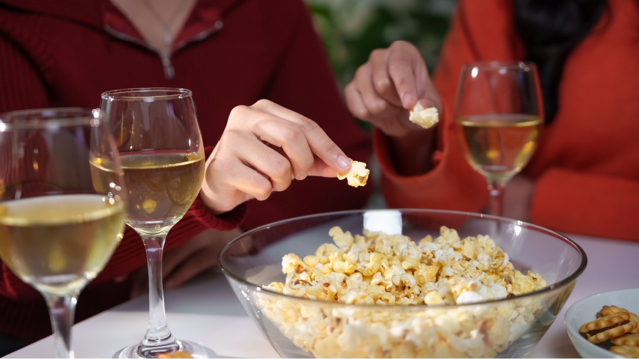 Two women are picking popcorn from a glass bowl while enjoying glasses of white wine, celebrating friendship and relaxation during a cozy home gathering