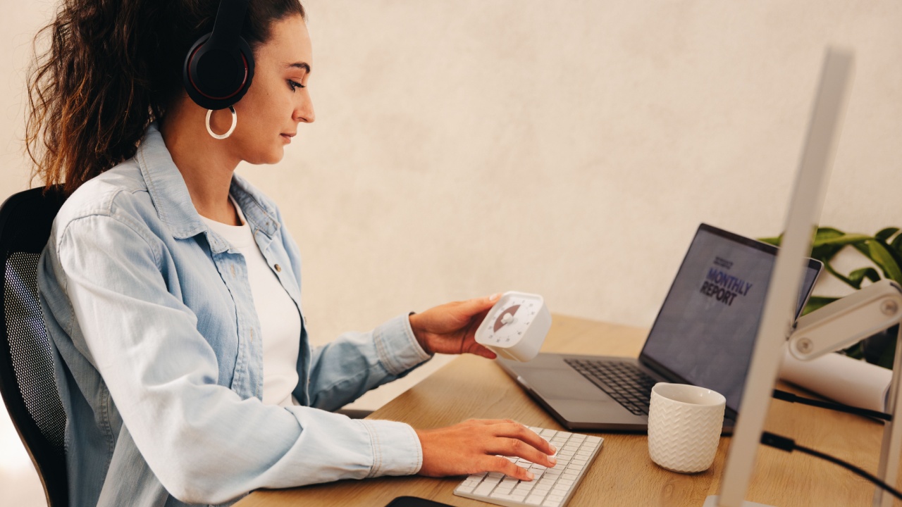 A woman wearing headphones working on her computer in an office. She holds a timer while staying focused on her tasks, showcasing productivity and attentive work attitudes.