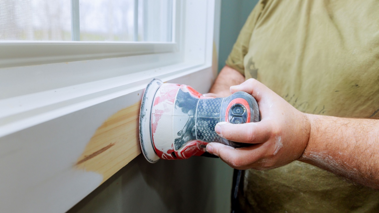 Wood worker is using an electric sander to smooth wooden trim by window, in renovation task.