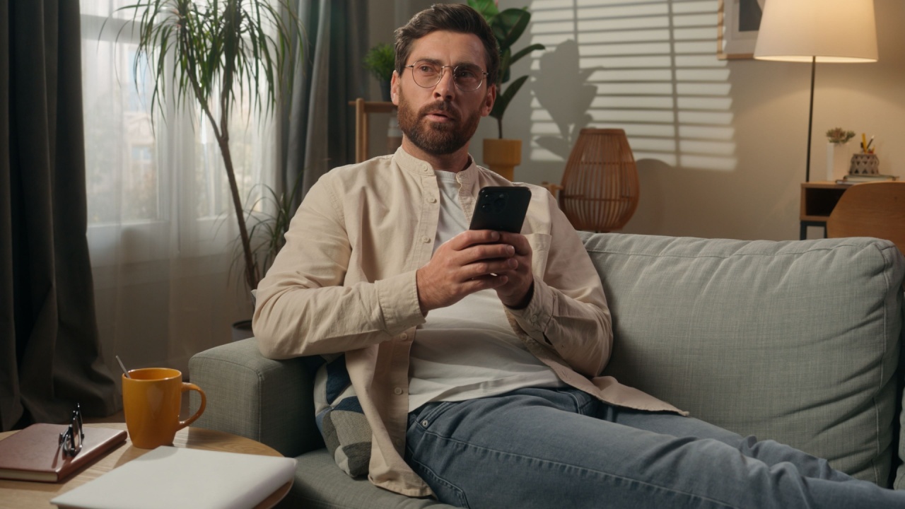 Man thoughtful dreaming Caucasian male homeowner tenant freelancer at home using mobile phone app online shopping in apartment smiling guy think on couch sofa relax browsing smartphone gadget addict
