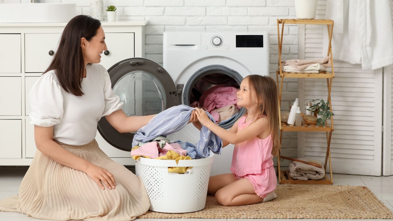 Young woman and her daughter pulling out fresh clothes from washing machine in laundry room