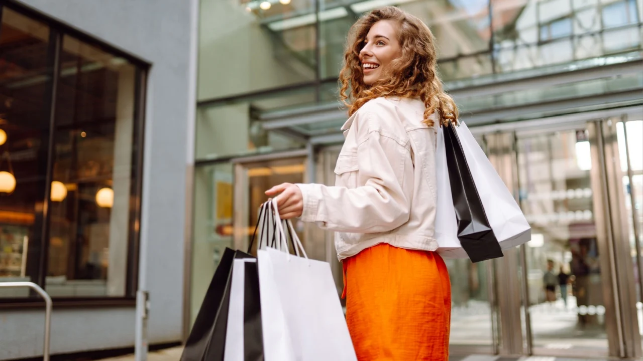 A joyful woman with shopping bags enjoys walking in a sunny city. Young woman after shopping outdoors. Consumerism, shopping concept.