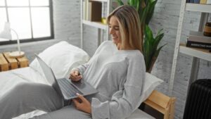 Woman relaxing on bed using laptop in cozy bedroom with plant decor and stylish gray outfit for comfortable indoor lounging and tech engagement.