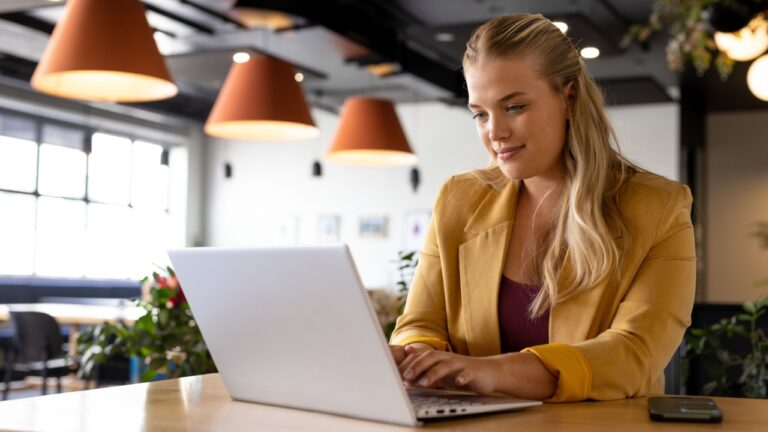 Young woman using laptop on table in coworking space, with smartphone and potted plants, copy space. Productivity, focus, remote, working, modern, minimalist, industrial