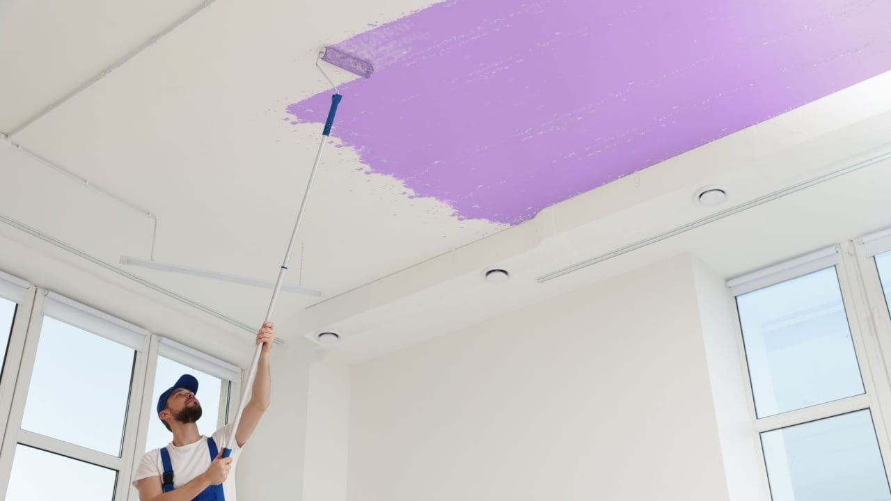 Man painting white ceiling with violet dye indoors, low angle view