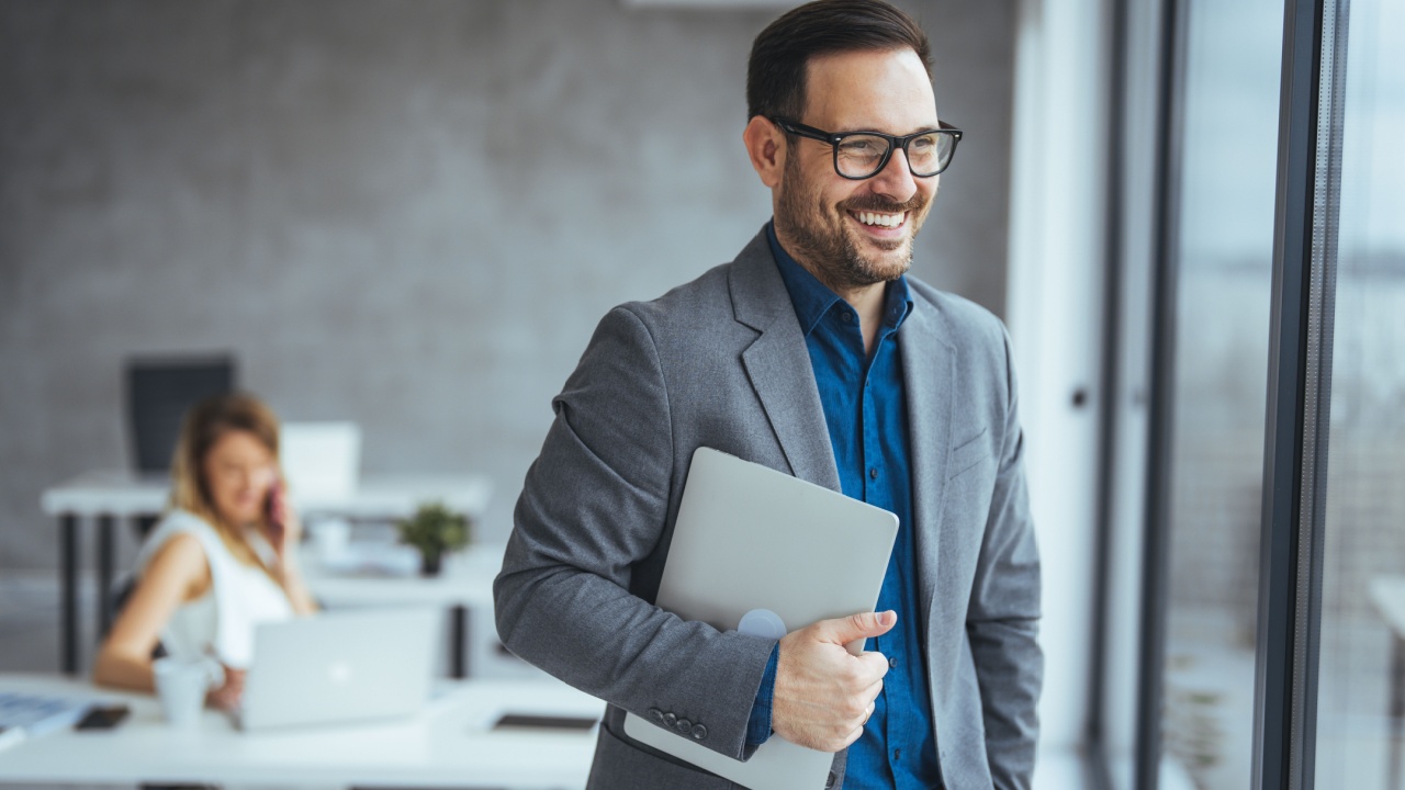 Professional executive standing confidently in a modern office, holding a laptop and smiling. Bright and airy working environment depicts a positive and productive workplace atmosphere.
