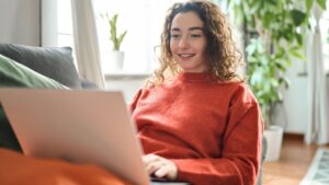 Young lady looking at computer ordering sale products on website, watching videos or elearning. Relaxed happy woman sitting on couch using laptop at home surfing, doing online ecommerce shopping,