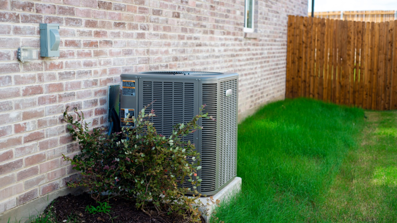 Outdoor HVAC unit and wall mounted electrical boxes installed alongside modern brick home, clean siding and windows, green grass landscaping shrub, privacy fence, disconnect switch, utility meter. USA