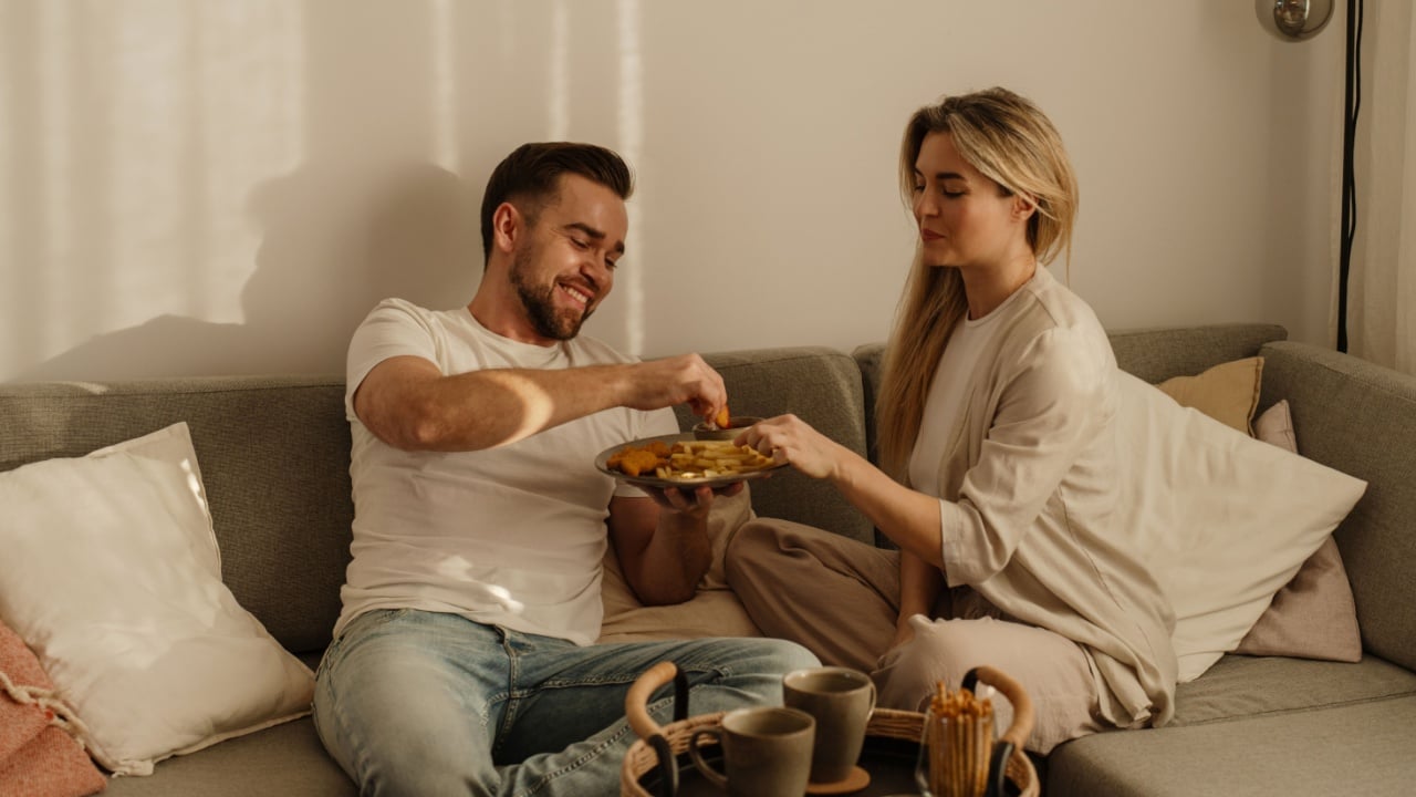 Couple sharing a plate of French fries and chicken nuggets in cozy lving room.