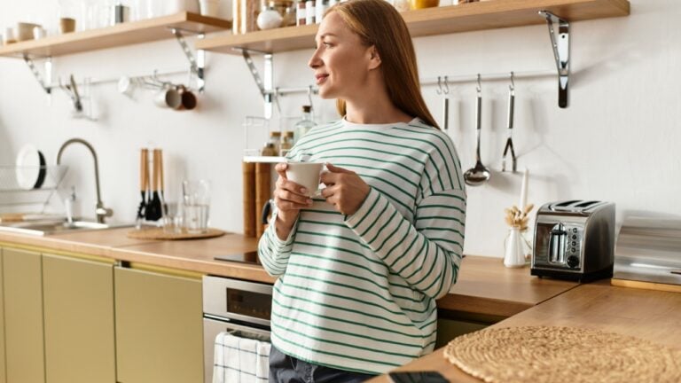 A beautiful young woman stands in her cozy kitchen, savoring a warm drink with a relaxed smile.