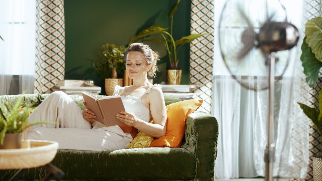 A woman with a gentle smile reclines on a green velvet sofa, eyes closed, holding an open book, lost in deep relaxation. A standing fan ensures comfort for her peaceful summer afternoon indoors.