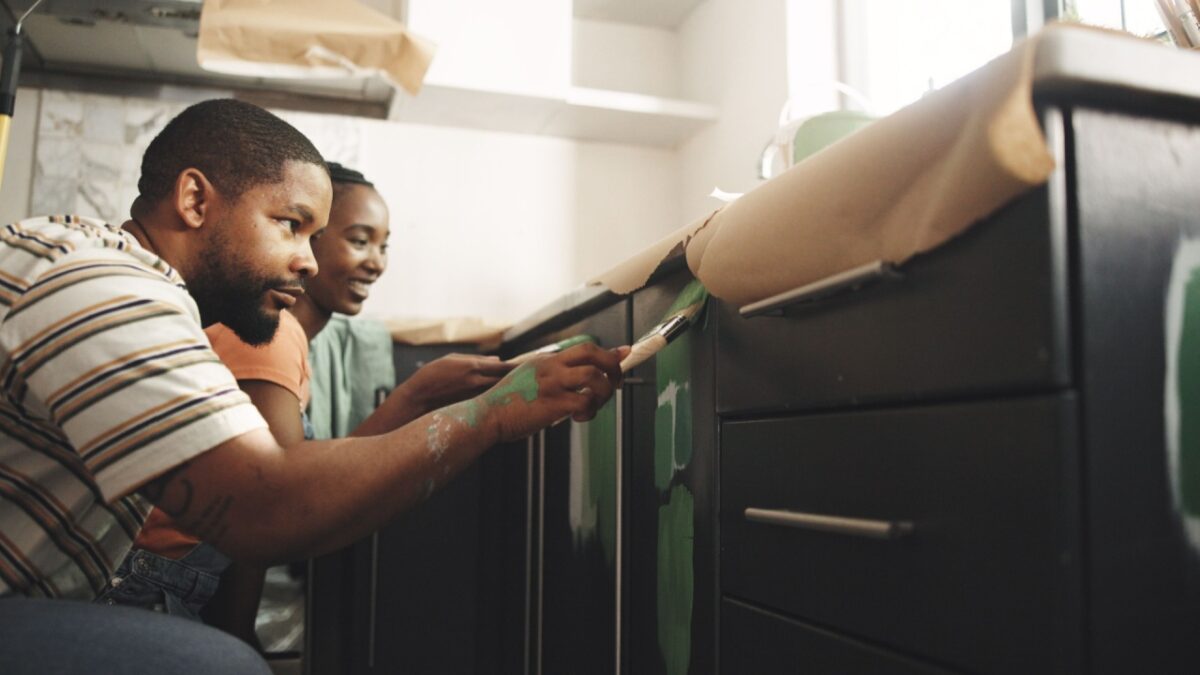 Painting, help and couple in home for improvement, diy project or remodeling together. African man, woman and paintbrush in house on kitchen cabinet for property renovation, creative color and smile