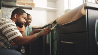 Painting, help and couple in home for improvement, diy project or remodeling together. African man, woman and paintbrush in house on kitchen cabinet for property renovation, creative color and smile