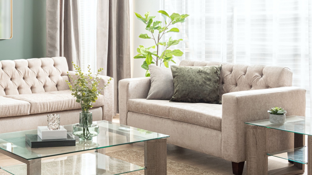Living Room Interior with Beige Fabric Loveseat Sofa Set and Rectangle Glass Coffee Table with Shelf and Wooden Legs. The Room is Illuminated by a Window with Beige Linen Curtain, Light Green Walls.