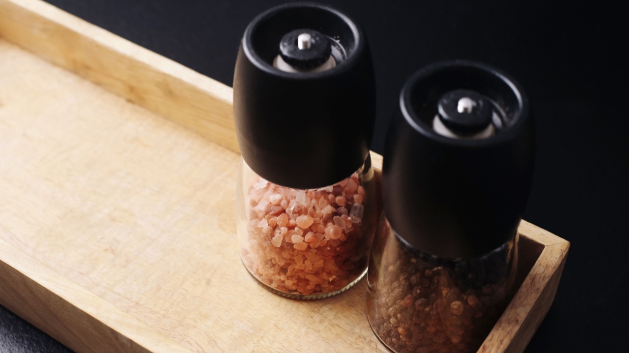 Pepper and salt grinders closeup. Kitchen seasoning set on wooden tray. Modern spice containers on black table. Culinary accessory for cooking and serving. Simple stylish home decor utensils.
