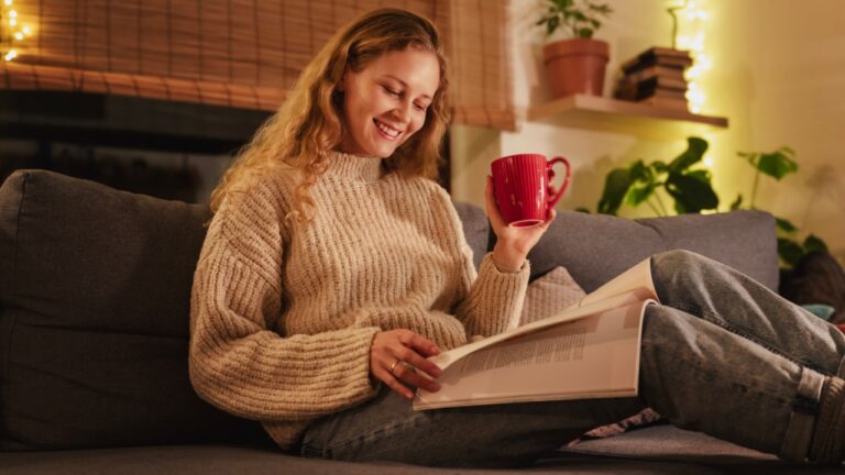 Young pretty curly young blonde woman lying on sofa at home reading magazine, cozy Christmas holidays