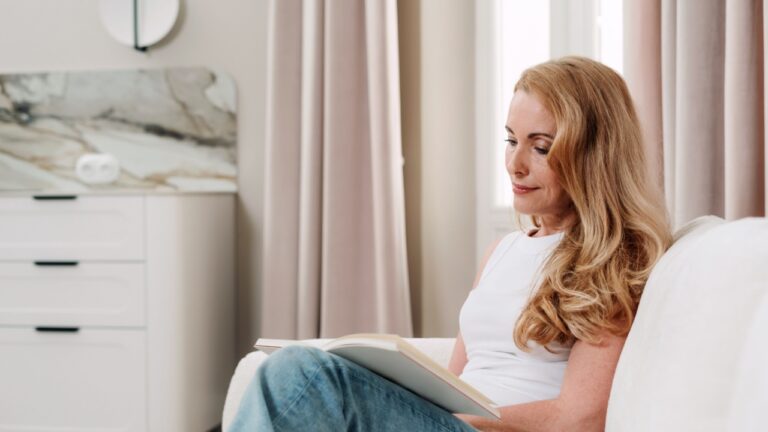 A mature woman relaxes alone in her comfortable living room, reeading a bestseller book. Soft curtains and minimalistic decor create a cozy atmosphere perfect for mindfulness moments on weekend