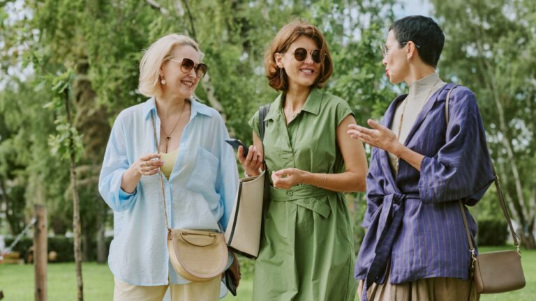 Three middle aged Caucasian women walking outdoors in park, smiling and talking together, holding handbags and wearing sunglasses, enjoying conversation in green natural setting