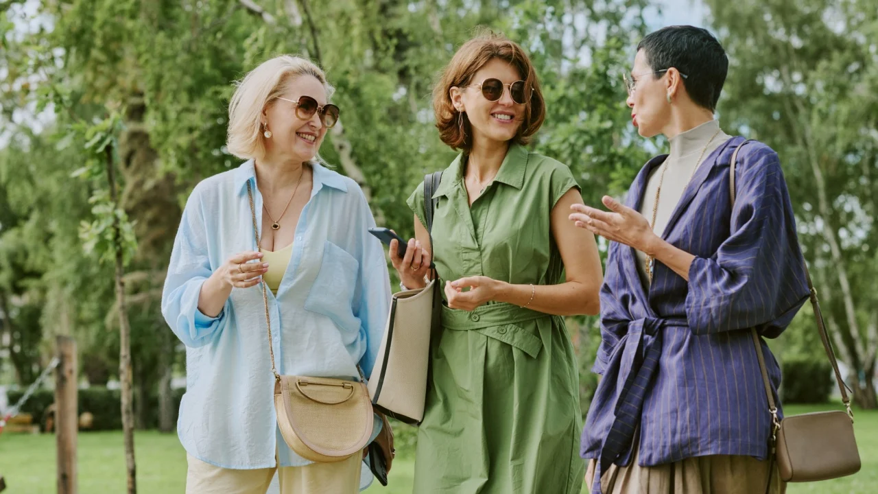 Three middle aged Caucasian women walking outdoors in park, smiling and talking together, holding handbags and wearing sunglasses, enjoying conversation in green natural setting