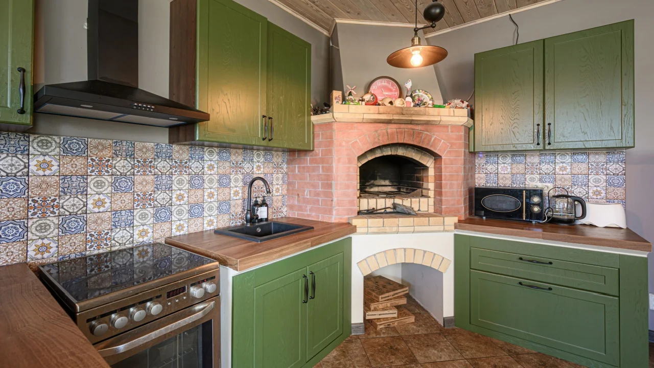 cozy kitchen with olive green cabinets, colorful backsplash tiles, and a brick oven. A modern stove sits beside the sink. Warm lighting illuminates the space