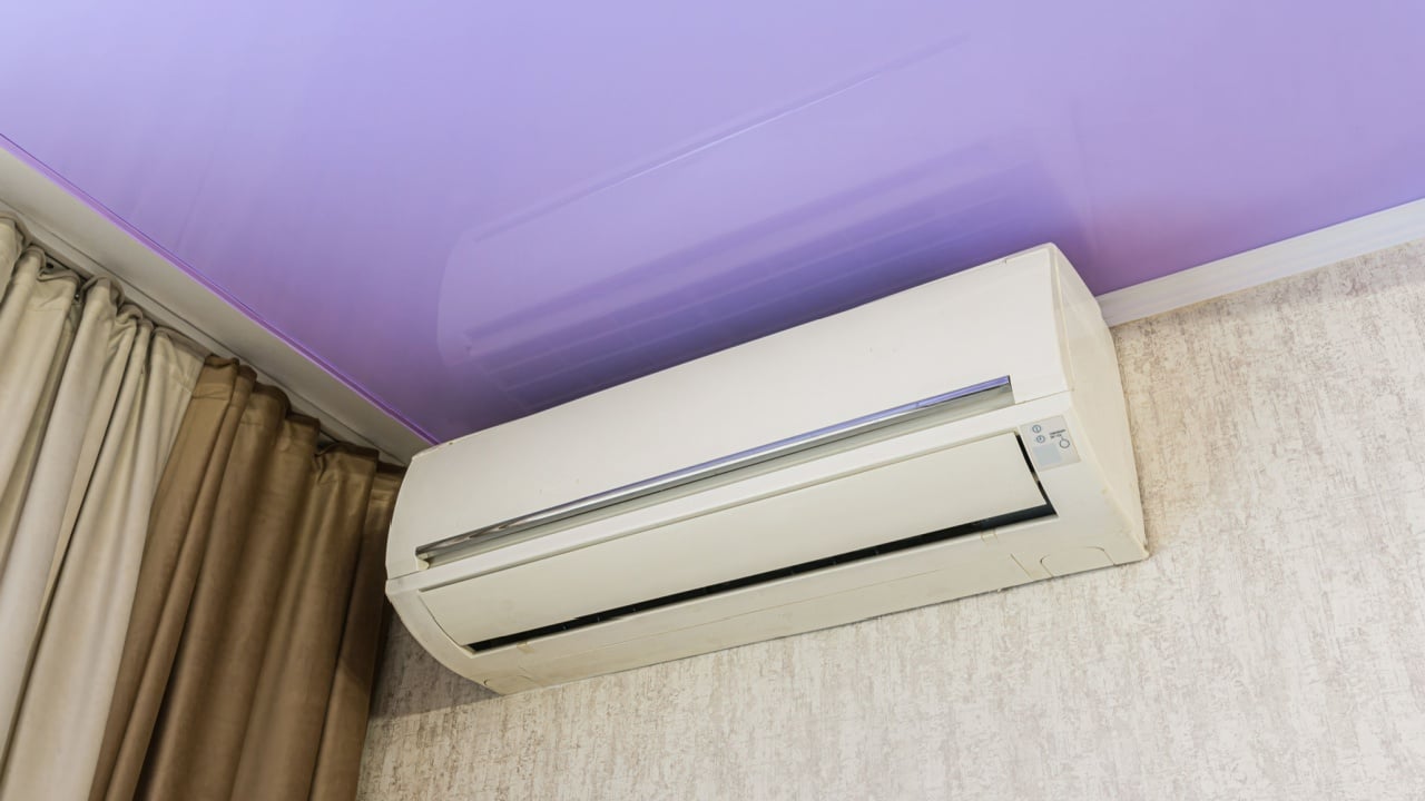 A white air conditioner unit is mounted on a textured wall, with curtains to the side and a purple ceiling above