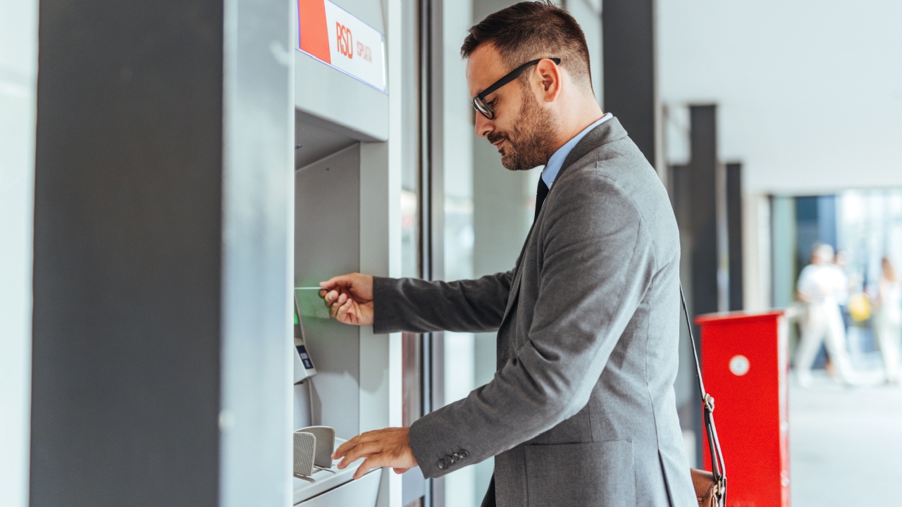A businessman wearing a suit and sunglasses withdrawing money from an ATM outdoors, symbolizing financial security, personal independence, and modern banking solutions.