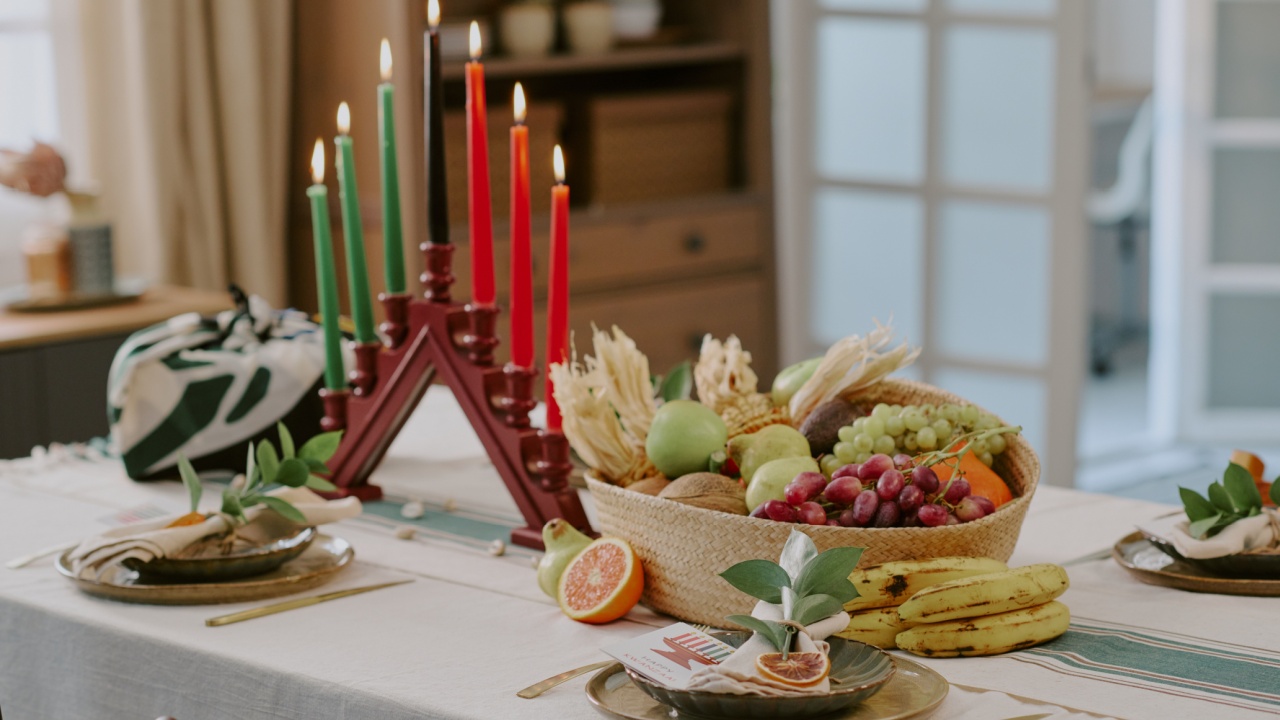 Kwanzaa kinara with seven lit candles standing on dining table surrounded by basket of assorted fruits and vegetables, traditional holiday setting celebrating heritage