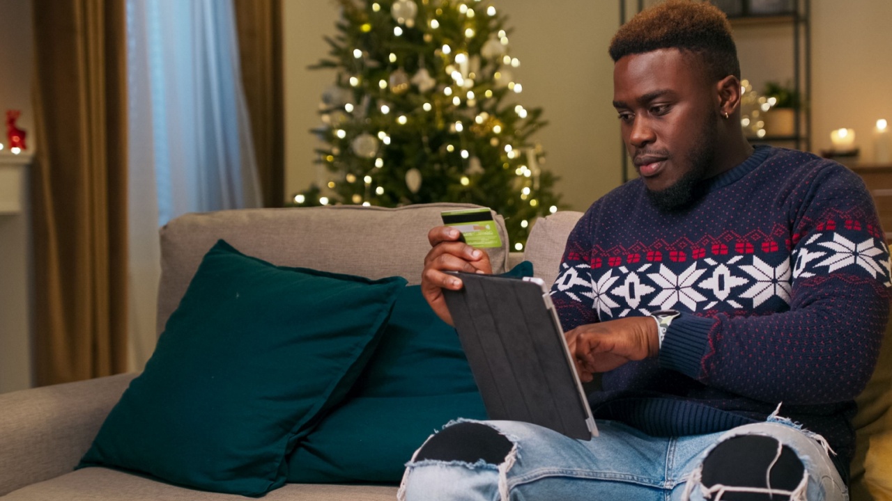 Holding a tablet and bank card, a young man of multi-racial background sits on a couch, ordering Christmas presents for friends. He carefully enters the card details. Dark blue and yellow pillows are