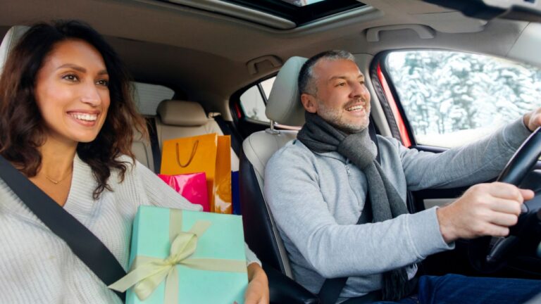 Happy European spouses driving car after shopping Xmas presents, woman holding wrapped gift box, shopper bags on back seat, couple preparing for New Year and Christmas