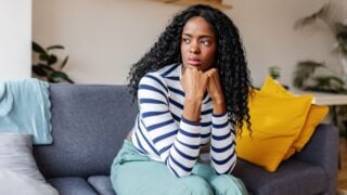 Young woman sits on a sofa at home, looking concerned and sad, reflecting on her mental health and facing challenges