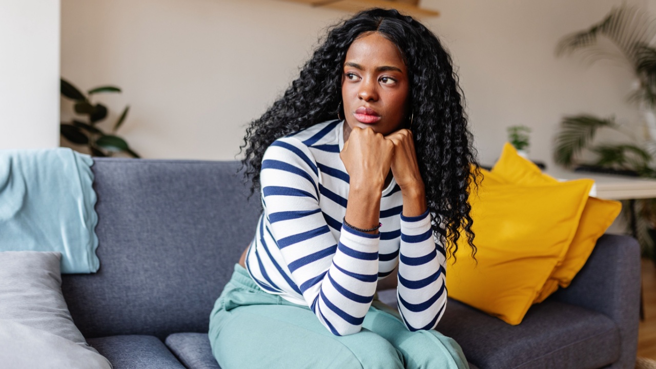 Young woman sits on a sofa at home, looking concerned and sad, reflecting on her mental health and facing challenges