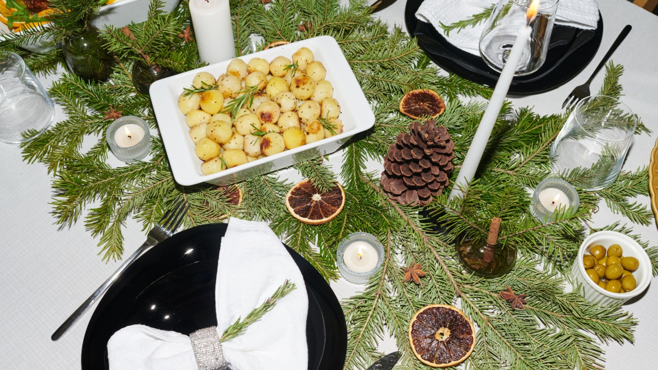 Christmas table setting featuring evergreen branches, pine cone, dried citrus slices, candles, black plates with napkins, bowl of roasted potatoes and dish of green olives