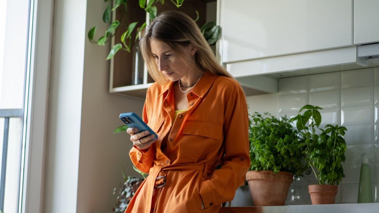 Relaxed woman holding phone, standing in cozy kitchen near window, leaning on counter with house plants. Scandinavian female scrolling social media on smartphone, reading news online, watching media.