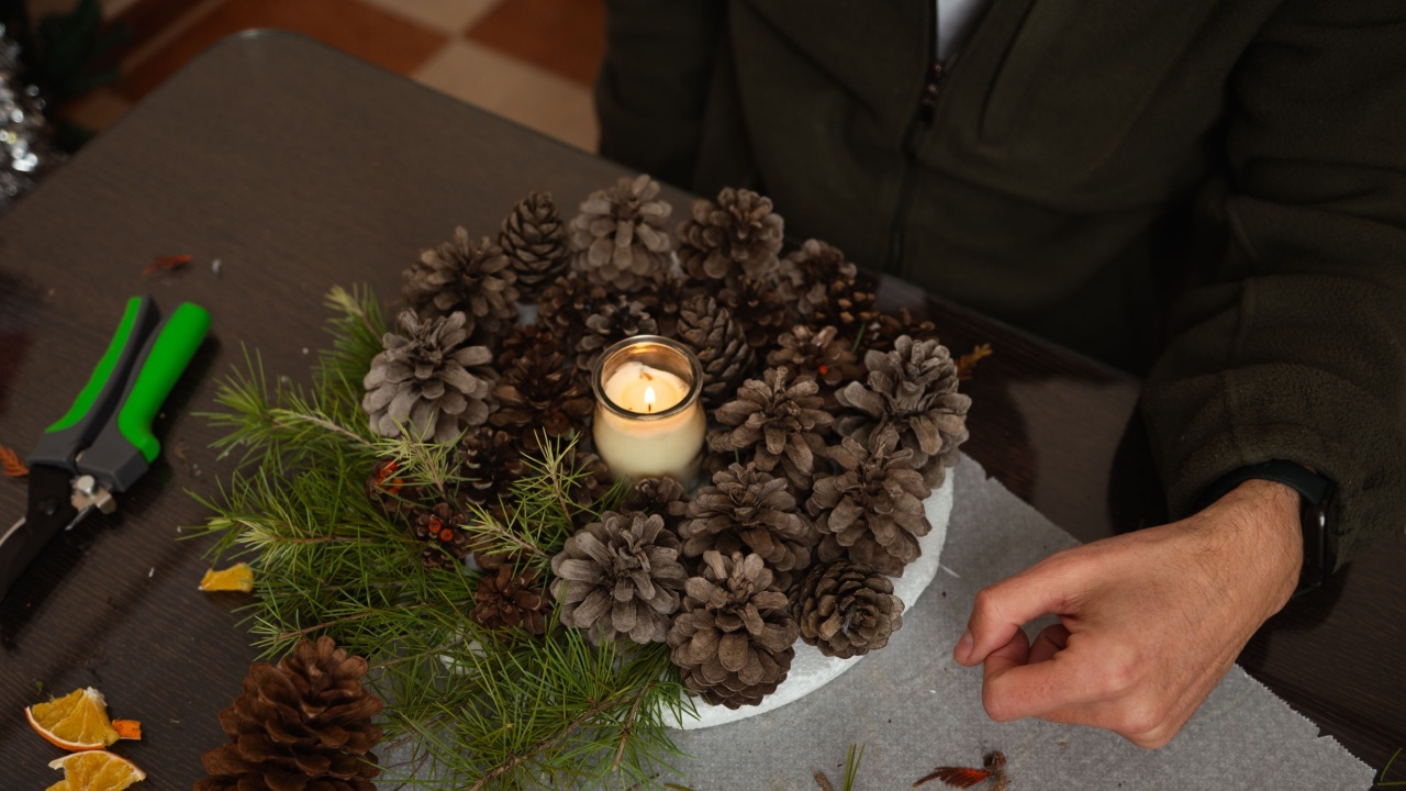 A cozy home scene of a family crafting Christmas decorations on a table, featuring pinecones, greenery, dried citrus, and a glowing candle centerpiece.