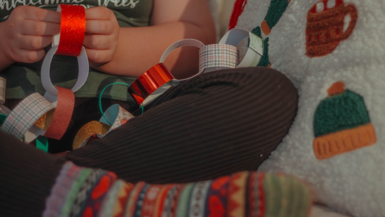 Person sitting on a coach wearing Christmas socks, making a festive paper garland, celebrating the holiday season.