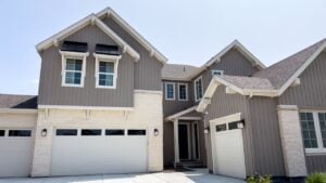 A large suburban house with stone and siding finishes stands on a clean concrete driveway. The bright day lighting emphasizes the crisp architectural lines and manicured landscaping.