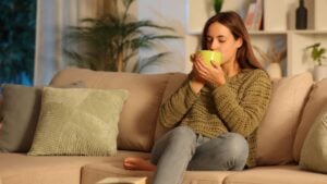 Woman in green drinking coffee or tea in the night sitting on a couch at home