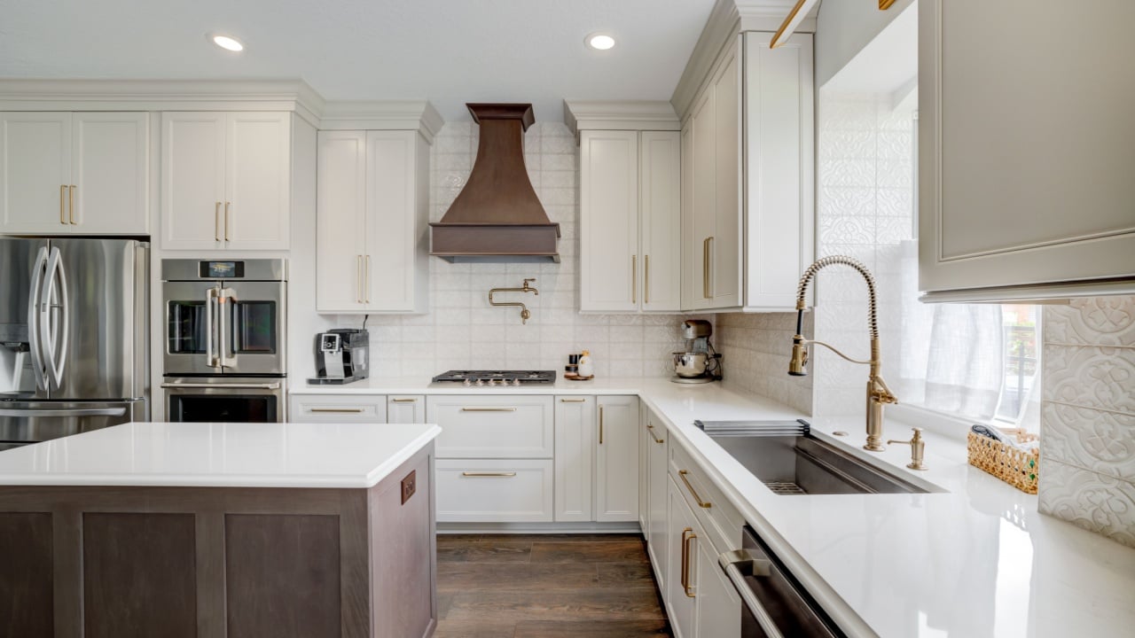 Modern luxury kitchen interior featuring white shaker cabinets, a large quartz island, stainless steel appliances, and a striking dark range hood. The space is bright and elegant with under-cabinet li
