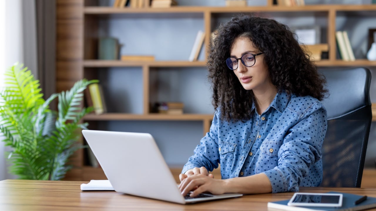 Young woman with curly hair and glasses concentrating on her work, typing on a laptop at a wooden desk in a modern home office with a bookshelf in the background