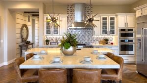 Bright contemporary kitchen with a large wooden dining table and leather chairs under pendant lighting. The white cabinets, stainless steel appliances, and patterned backsplash create a sophisticated