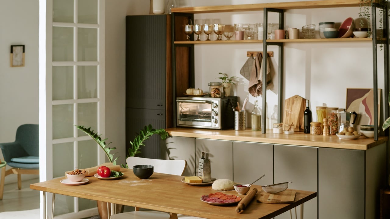 Modern kitchen interior featuring wooden table with various ingredients and utensils arranged for meal preparation, open shelving displaying glassware and kitchen accessories