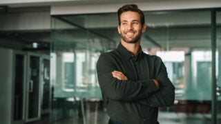 Corporate portrait of European confident student, trade manager businessman standing crossed arms in office. Young latin hispanic specialist business man employee smiling dreaming away.