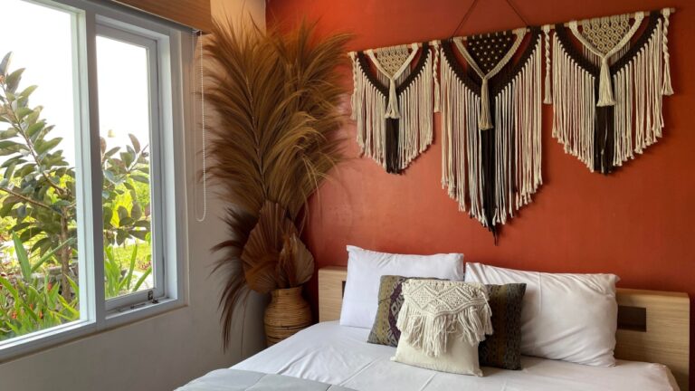 A cozy and stylish bedroom featuring a warm red accent wall decorated with intricate macramé wall hangings. The bed is neatly made with white linens and a grey throw, flanked by a large dried pampas grass arrangement in a woven vase near a bright window.