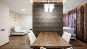 A loft-style dining area with a wooden table, white chairs, dark brick accent wall, modern pendant light, plush sofas, and textured curtains