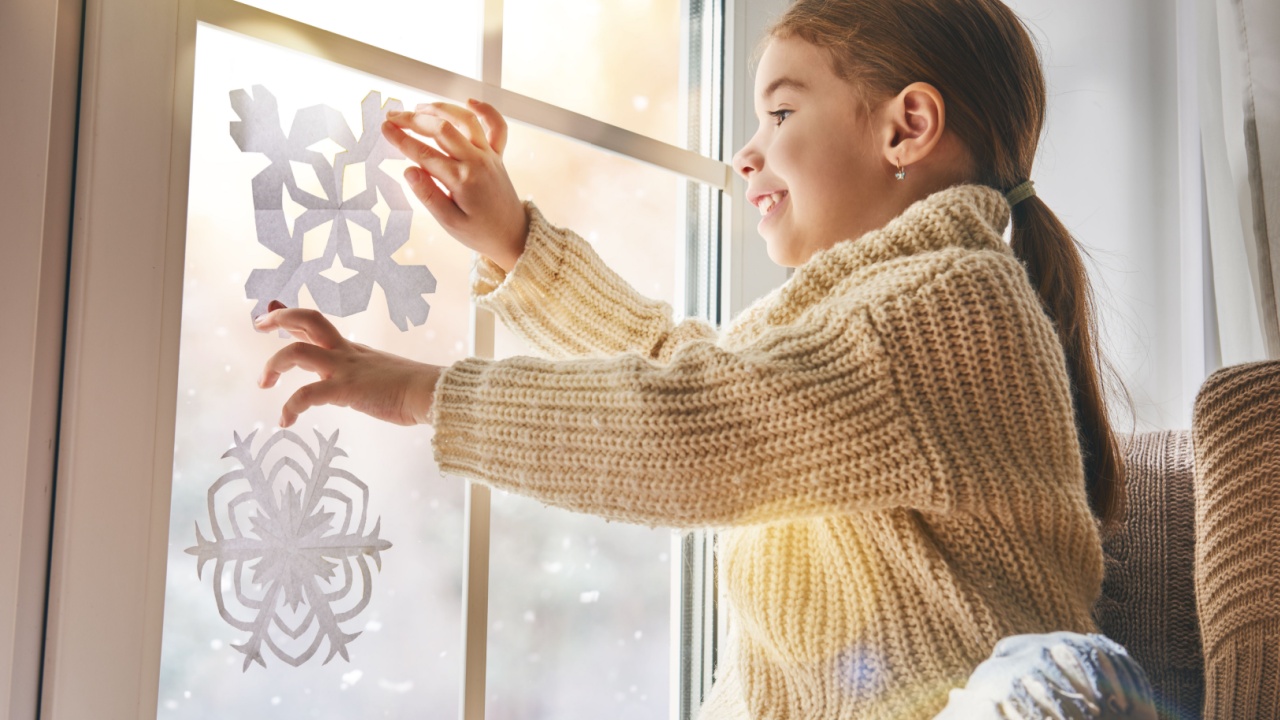 Cute little girl sitting by the window and looking at the winter forest. Child makes paper snowflakes for decoration windows. Kid enjoys the snowfall.