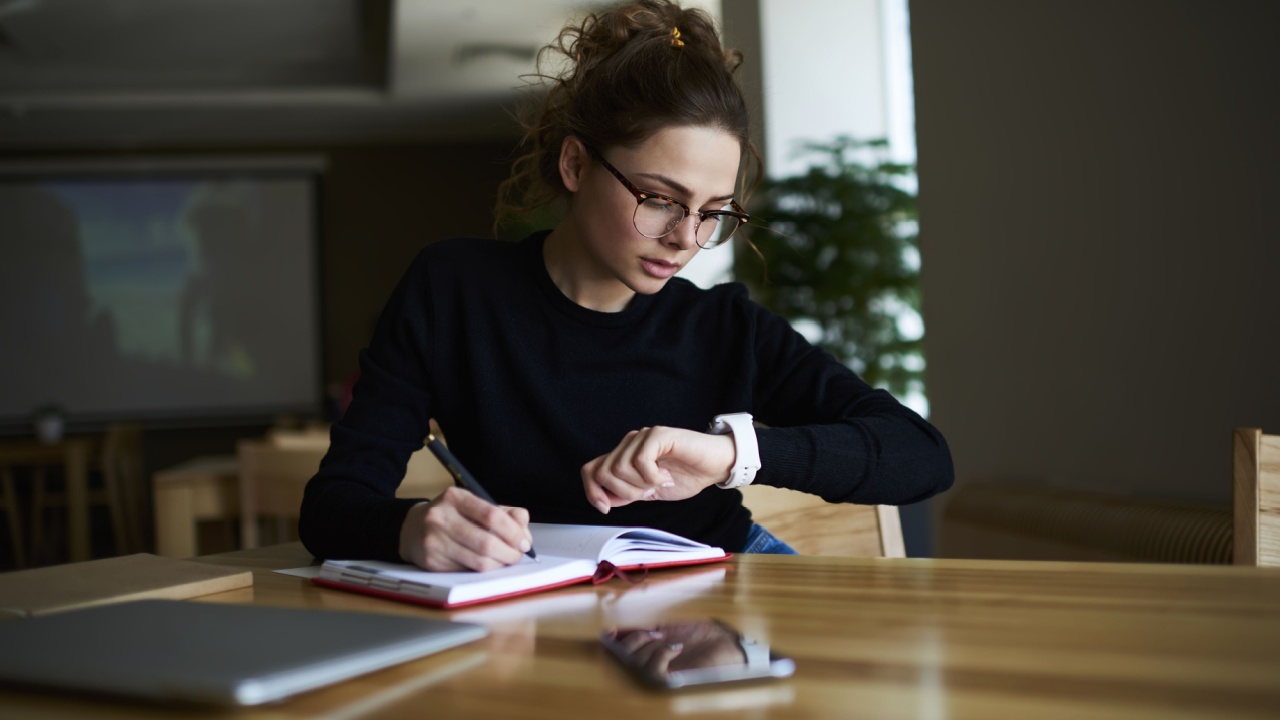 Talented female writer in stylish eyeglasses noting some information in notepad while looking on alarm watch to managing time for organization of working process sitting in university indoors