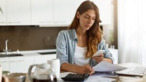 Candid shot of beautiful concentrated young Caucasian woman typing text message on mobile phone while calculating bills in kitchen, sitting at table with laptop computer, papers and calculator