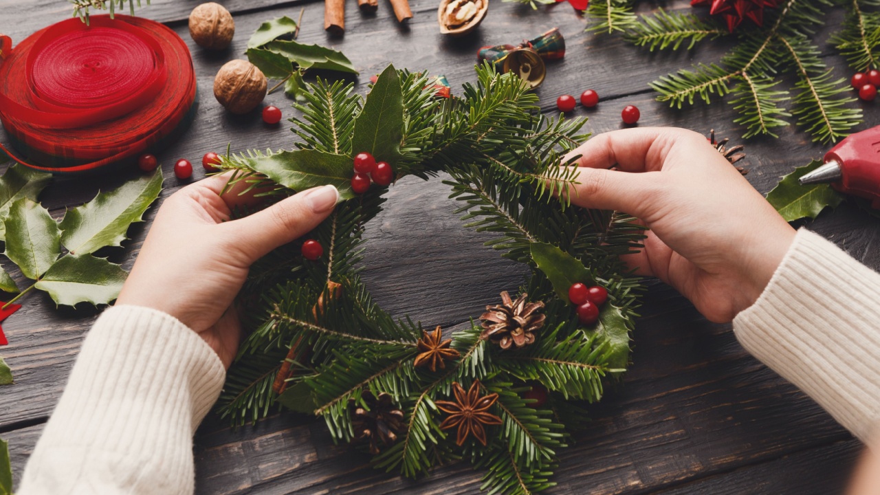 Point of view of wooden table background with female hands making wreath. Creative leisure, cup, tools and trinkets for xmas holiday decoration.