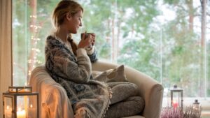 Young beautiful woman sitting home in the chair by the window with cup of hot coffee wearing knitted warm sweater. Cozy room decorated with lanterns and candles. Stay home